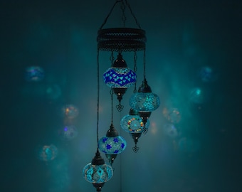 May include: A hanging chandelier with six mosaic glass lamps in shades of blue and turquoise. The lamps are suspended from a decorative metal frame and chain. The lamps are illuminated, creating a warm, inviting glow.