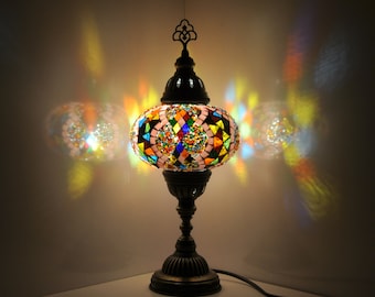 Puede incluir: Una lámpara de mesa de mosaico de vidrio de colores con una pantalla redonda con patrones rosa, azul, verde y amarillo. La lámpara tiene una base de bronce y un cable.