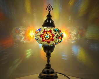 Puede incluir: Una lámpara de mesa de mosaico colorida con un diseño floral. La lámpara tiene una base redonda y un tallo alto y delgado. Los azulejos de mosaico son en tonos de rojo, naranja, amarillo, verde, azul y blanco.