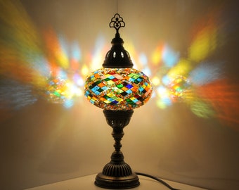 Puede incluir: Lámpara de mesa decorativa con pantalla de mosaico de vidrio. La pantalla está compuesta por piezas de vidrio de colores en forma de diamante. La lámpara tiene una base y un tallo de metal oscuro, y la luz proyecta patrones coloridos en la pared.