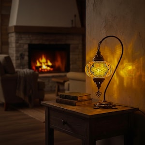 May include: A decorative table lamp with a mosaic glass shade in shades of yellow and orange. The lamp has a curved, dark metal frame and sits on a wooden nightstand. Two books are stacked on the table, and a fireplace is in the background.