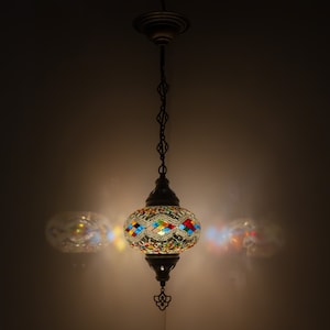 Może przedstawiać: Wisząca lampa z okrągłym, kolorowym abażurem z mozaiki szklanej. Abażur ma geometryczny wzór z niebieskich, zielonych, czerwonych, żółtych i białych płytek. Lampa jest zawieszona na łańcuchu i ma metalową podstawę.