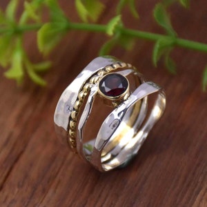 May include: A silver and gold ring with a red gemstone. The ring is made of three bands, one of which is textured with small beads. The ring is set against a wooden background.