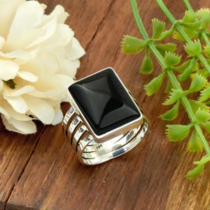 Può includere: Anello d'argento con una grande pietra di onice nera rettangolare. L'anello ha un design a più bande ed è posto su una superficie di legno, con accenti di fiori bianchi e piante verdi.