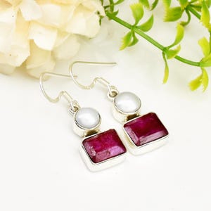 Könnte beinhalten: Silberne Ohrhänger mit einer weißen Perle und einem roten Rubin-Edelstein. Die Ohrringe haben ein schlichtes Design und sind perfekt für den Alltag.
