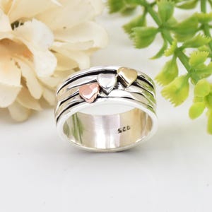 May include: A silver spinner ring with three heart accents in rose gold, silver, and gold. The ring has a textured band and the number "925" is stamped on the inside.