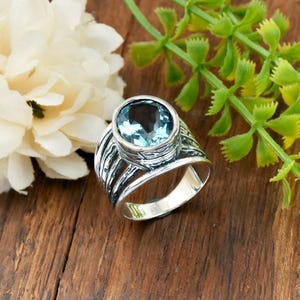 May include: A silver ring featuring a large, oval, pale blue gemstone. The ring has a textured band with vertical lines and a smooth interior. The ring is set against a wooden surface with a white flower and green foliage.