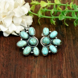 May include: A pair of stud earrings featuring multiple oval-shaped turquoise stones set in silver. Each earring has a central stone surrounded by smaller stones arranged in a fan-like pattern with twisted silver wire detailing.