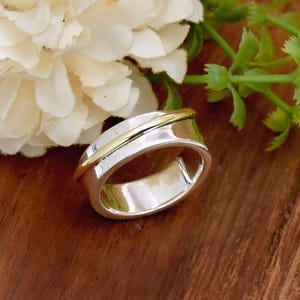 May include: A silver ring with a thin gold band wrapped around it. The ring is wide and has a smooth, rounded surface.