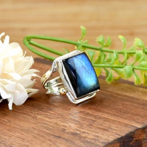 May include: A silver ring with a rectangular, iridescent blue gemstone. The ring has a gold accent on the band and a gold bead. The ring is displayed on a wooden surface with white flowers and green foliage in the background.