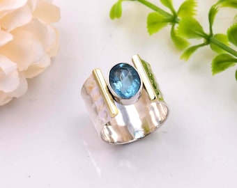 May include: A silver ring with a blue gemstone set in a gold band. The ring has a hammered texture and two gold bars on either side of the stone.