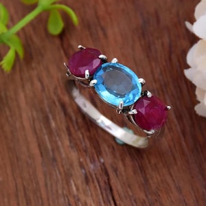 Può includere: Un anello d'argento con tre pietre ovali. La pietra centrale è un topazio blu chiaro, e le due pietre ai lati sono rubini rossi.