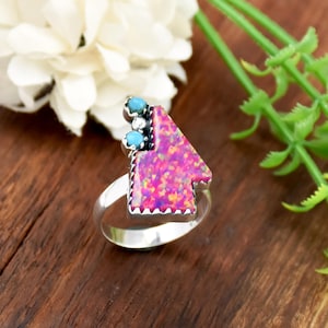 May include: A silver ring featuring a pink and purple arrow-shaped stone with turquoise accents. The ring has a detailed design and is set against a wooden surface, with white flowers and green foliage in the background.