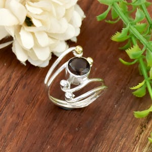 Può includere: Un anello d'argento con una pietra preziosa marrone scuro, accentuato da dettagli in oro e argento. L'anello presenta un design unico e aperto con fasce curve e piccole sfere decorative. La pietra preziosa è rotonda e incastonata in una lunetta d'argento.