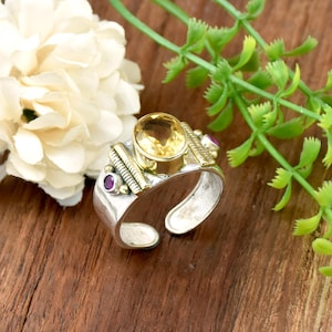 Peut inclure: Bague argentée ornée d'une grande pierre jaune ovale, agrémentée de petites pierres violettes et de détails en métal doré. La bague, de style ouvert, est présentée sur une surface en bois avec des éléments floraux.