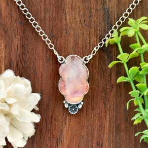 May include: A silver necklace featuring a pendant with a light pink and orange gemstone. The pendant has a unique, scalloped shape and is accented with a small floral design at the bottom. The chain is a delicate link style.
