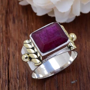 Può includere: Un anello in argento e oro con una grande pietra preziosa quadrata di colore rosso scuro. L'anello ha una fascia d'oro intrecciata e piccole sfere d'oro su entrambi i lati della pietra.