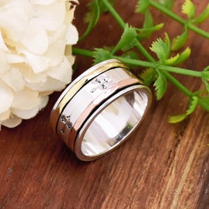 May include: A silver spinning ring with a cross design. The ring has three bands of gold and rose gold.