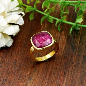May include: A gold-toned ring featuring a large, square-cut, faceted magenta gemstone set in a smooth metal bezel. The ring band is solid and polished, showcasing a vibrant pink stone with visible internal texture and light-reflecting facets.