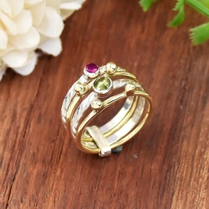 Könnte beinhalten: Ein stapelbares Ring-Set mit Silber- und Golddesign. Die Ringe weisen eine Kombination aus glatten Bändern und strukturierten Silberabschnitten auf. Die Ringe sind mit kleinen Goldkügelchen und zwei Edelsteinen verziert: einem runden grünen Stein und einem kleineren, ovalen rosa Stein.