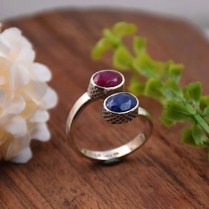 May include: A silver ring featuring two oval-shaped gemstones. The larger gemstone is a deep red ruby, and the smaller gemstone is a blue sapphire.
