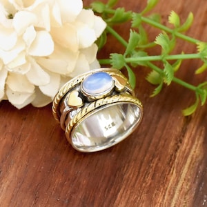 Puede incluir: Un anillo de plata y oro con una piedra de luna azul y acentos en forma de corazón. El anillo es un anillo giratorio con una banda que gira.
