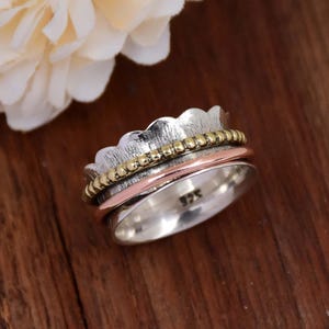 Può includere: Un anello girevole in argento e rame con una fascia testurizzata e una fila di perline dorate. L'anello è progettato per girare liberamente ed è una scelta popolare per la meditazione e il sollievo dall'ansia.