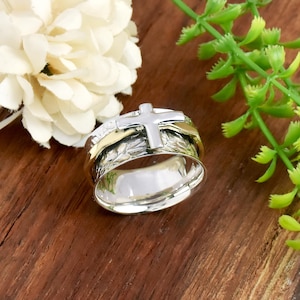 Peut inclure: Une bague argentée et dorée avec un motif de croix. La bague présente une croix argentée sur le dessus, avec des bandes dorées et une bande argentée texturée. La bague est posée sur une surface en bois avec de la verdure et une fleur blanche.