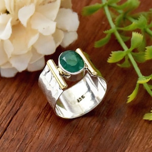 Könnte beinhalten: Ein silberner Ring mit gehämmerter Textur und goldenen Akzenten. Ein großer grüner Edelstein ist in der Mitte des Rings eingefasst.