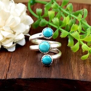 May include: A silver bypass ring featuring three round, polished turquoise cabochons set in scalloped bezels. The band wraps around the finger in a spiral design, showcasing the bright blue-green stones against the smooth, reflective silver metal.