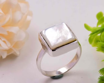 May include: A silver ring featuring a square, iridescent mother-of-pearl stone. The ring has a simple band and a bezel setting that holds the pearl. The ring is shown against a soft, blurred background with hints of green and cream.