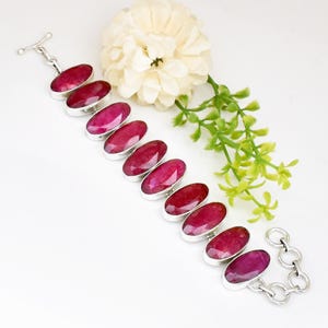 May include: A silver bracelet featuring oval-shaped, deep red gemstones. The gemstones are set in individual silver bezels and connected by silver links. The bracelet is displayed on a white surface with a white flower and green sprigs.