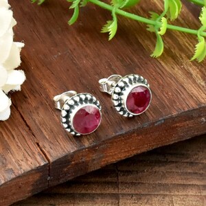 May include: A pair of stud earrings featuring round, faceted deep red gemstones set in textured silver bezels with a beaded border. The earrings have standard post backs and are displayed as a matching set, showcasing the vibrant red stone color.
