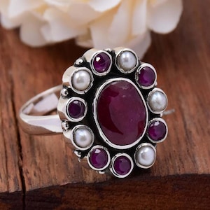 May include: A silver ring with a large oval-shaped red gemstone surrounded by smaller red gemstones and white pearls.