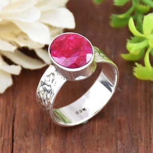 May include: A silver ring with a bright red oval gemstone. The band of the ring has a textured design.