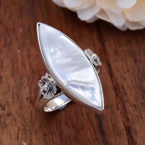 May include: A silver ring with a white mother-of-pearl oval stone and floral accents.
