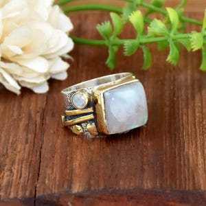 May include: A silver and gold-toned ring featuring a large, square, pale blue gemstone. The ring has a textured band with gold accents and a smaller, round, pale blue stone. The ring is set on a wooden surface.