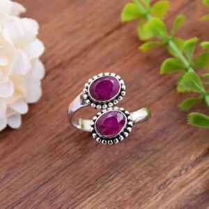 Puede incluir: Un anillo de plata con dos piedras preciosas ovaladas de color rojo intenso. Las piedras preciosas están rodeadas por un borde de cuentas. El anillo está sobre una superficie de madera, con una flor blanca y hojas verdes en el fondo.