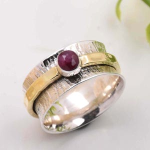 May include: A silver and gold spinning ring with a red gemstone. The ring has a textured band and a smooth, polished inner band.