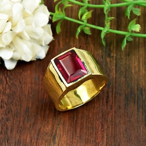 Könnte beinhalten: Ein goldener Ring mit einem großen, rechteckigen, tiefroten Edelstein in der Mitte. Die Ringschiene ist breit und poliert mit einer glatten, reflektierenden Oberfläche. Der Edelstein hat einen Facettenschliff, der das Licht einfängt.