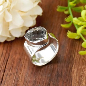 Peut inclure: Une bague en argent ornée d'une grande pierre brute gris foncé montée sur un anneau en métal poli. L'anneau présente un design ouvert et ajustable avec une surface lisse et réfléchissante qui contraste avec l'aspect texturé de la pierre.