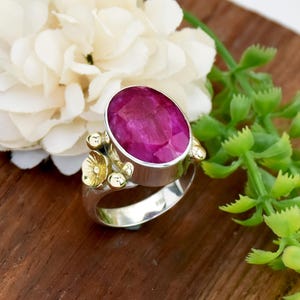 May include: A silver ring with a large, oval-cut ruby gemstone. The ring has gold floral accents on the sides. The ruby is a deep pink colour, and the ring is set against a wooden surface with white flowers and green foliage.