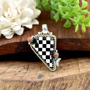 Può includere: Ciondolo in argento con motivo a scacchi bianchi e neri. Il ciondolo ha una forma triangolare con bordo dorato e due piccole pietre color corallo. Appoggiato su una superficie di legno.