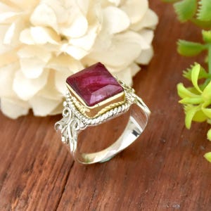 May include: A silver ring with a square ruby gemstone set in a gold bezel. The ring has a detailed, ornate design.