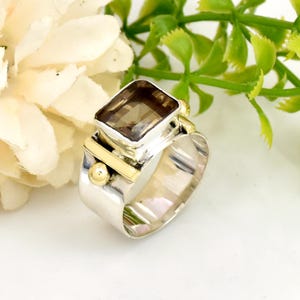 Könnte beinhalten: Ein Silber- und Goldring mit einem quadratischen, rauchquarzfarbenen Edelstein. Der Ring hat ein breites Band mit einem strukturierten Design.
