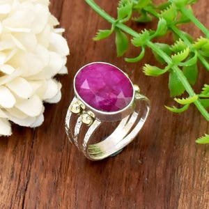 May include: A silver ring featuring a large, oval-shaped, faceted magenta gemstone set in a bezel. The band is composed of three parallel silver wires with textured detailing and two small, round, gold-toned accents on each side of the setting.