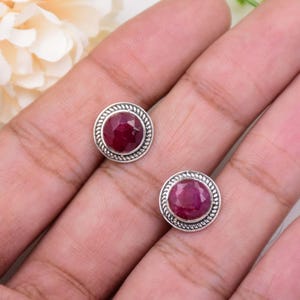 Può includere: Un paio di orecchini a lobo rotondi con pietre preziose di rubino rosso scuro sfaccettate incastonate in montature d'argento lucido. Ogni orecchino è circondato da un bordo decorativo a corda intrecciata che crea un profilo metallico testurizzato.