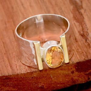 Peut inclure: Une bague en argent et or avec une grande pierre précieuse orange de forme ovale sertie au centre. La bague a une large bande avec une finition texturée.