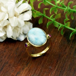 May include: A gold ring featuring a large, oval-shaped, light blue Larimar gemstone in the center, flanked by two small, round, deep pink rubies set on either side of the band. The metal band has a polished, reflective gold finish.