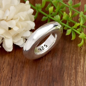Puede incluir: Un anillo de plata pulida con una banda gruesa y redondeada. El interior de la banda está claramente marcado con el sello 925, que indica la composición del metal. El anillo tiene una superficie lisa y reflectante con un aspecto sólido.